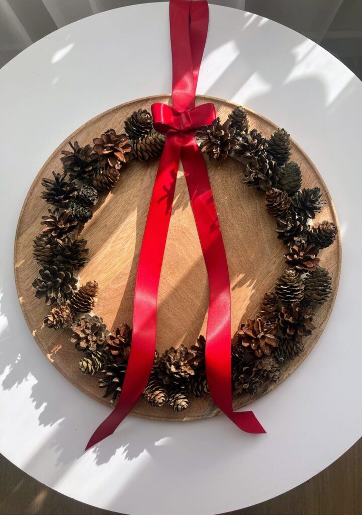 Ręcznie robiony wieniec świąteczny z naturalnych szyszek na drewnianej bazie, ozdobiony czerwoną satynową wstążką i kokardą. Rustykalna, bożonarodzeniowa dekoracja w stylu skandynawskim, idealna na drzwi, ścianę lub nad kominek.