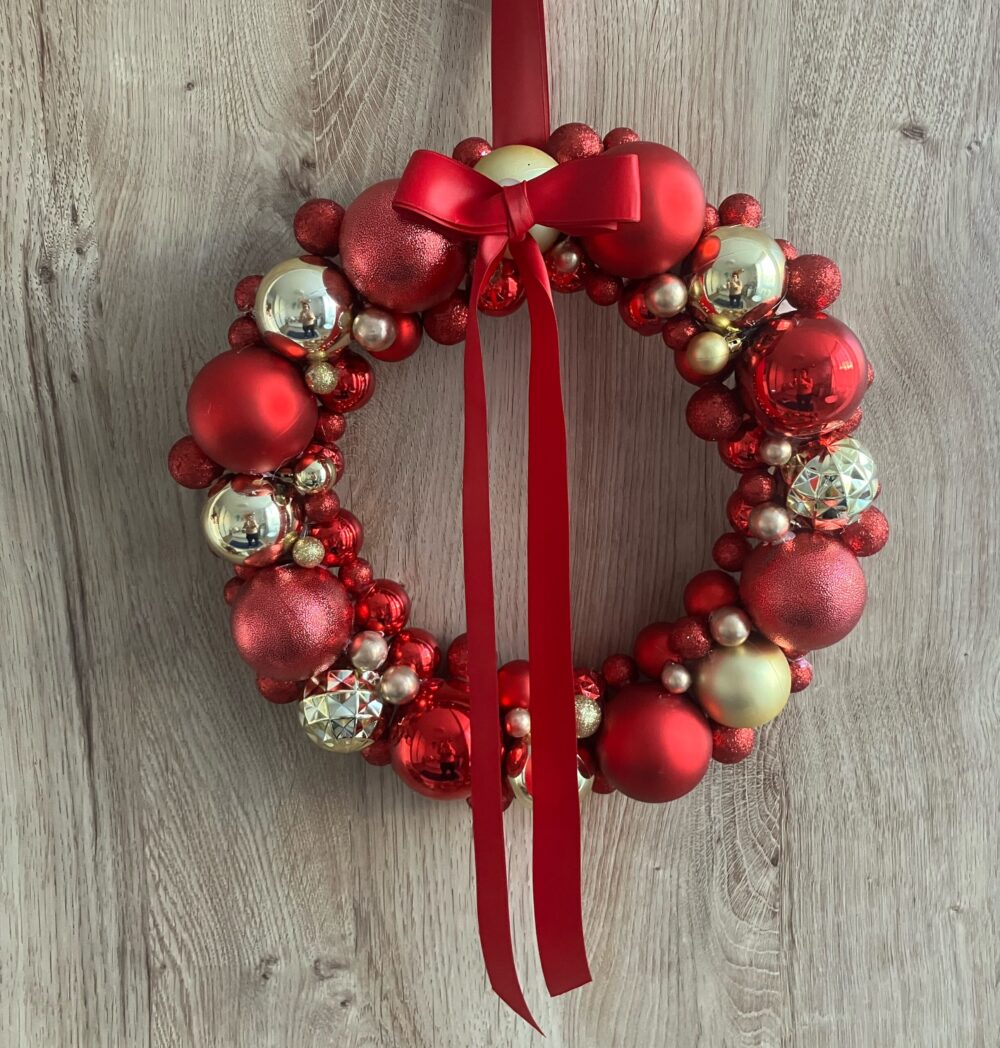 Czerwono-złoty wianek bożonarodzeniowy z bombkami i satynową czerwoną kokardą na drewnianej podstawie.