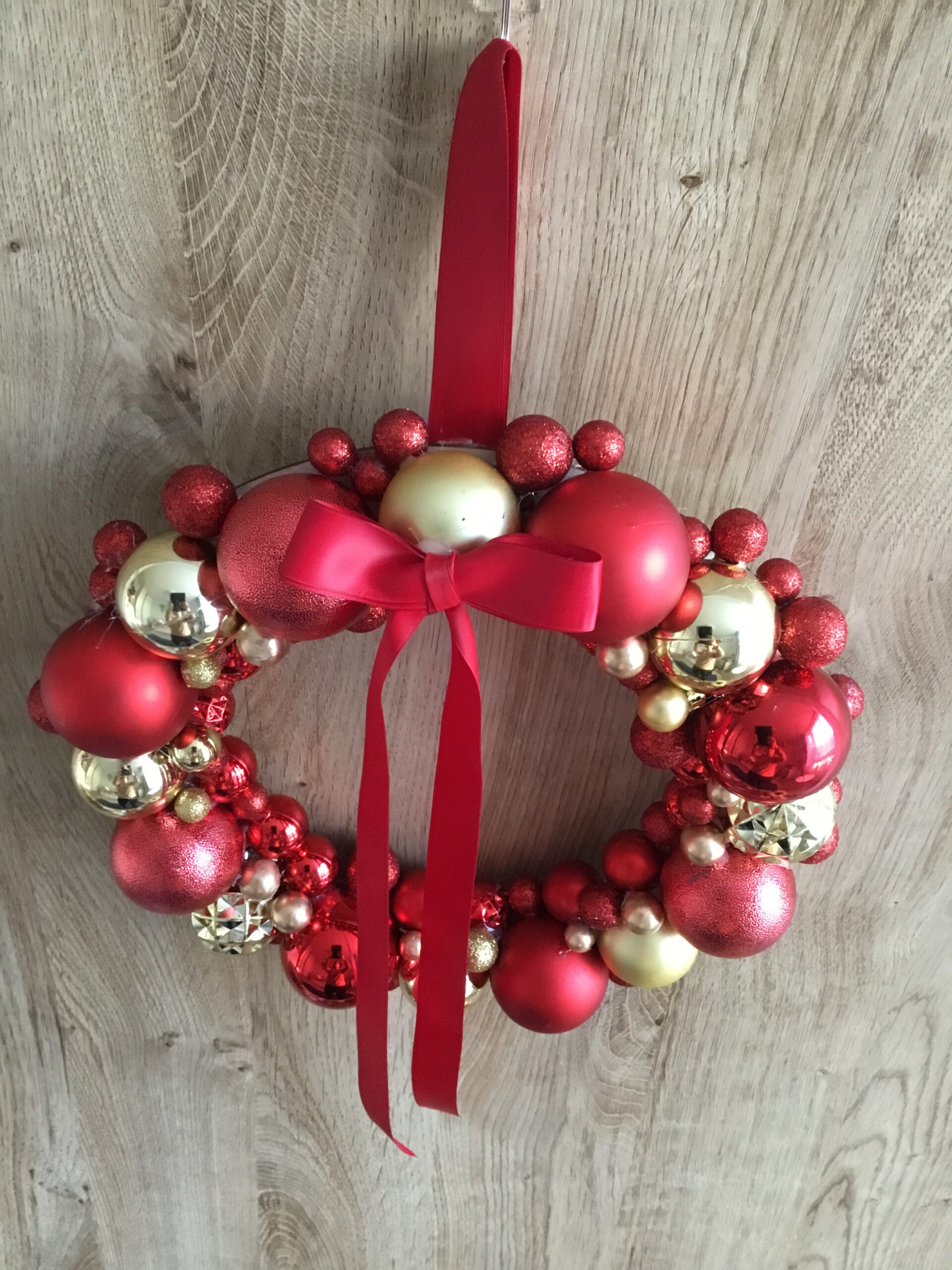 Świąteczny wianek o średnicy 30 cm, ozdobiony bombkami i czerwoną satynową wstążką, zawieszony na drzwiach.